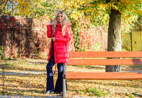 Lady attractive fashionista posing in jacket. Woman fashionable blonde with makeup stand in autumnal park. Jacket for fall season concept. Girl wear red bright warm jacket. Fall fashion concept