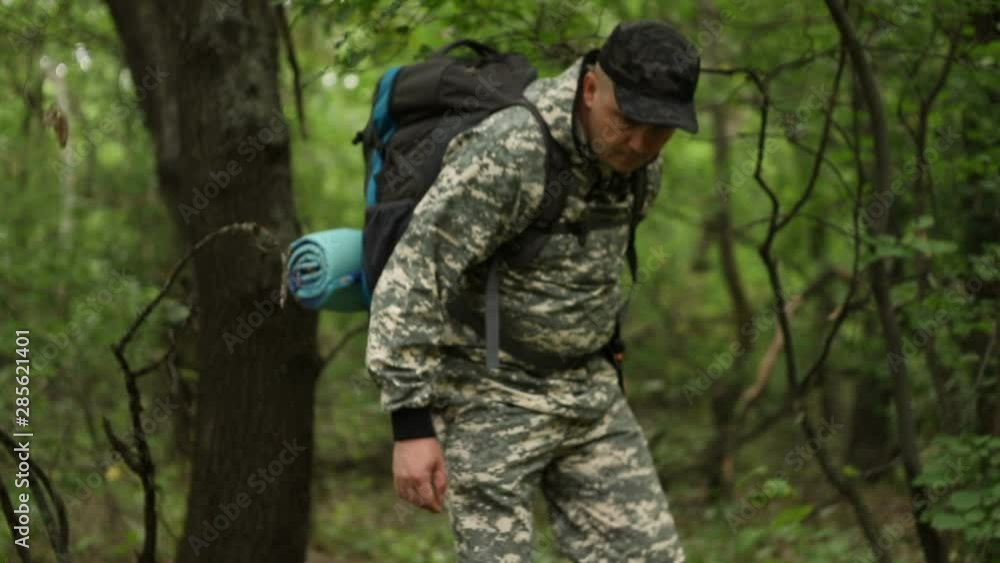 young handsome сaucasian man in a camouflage suit, cap and with a backpack, walks through the dense green forest past the camera, the average plan young handsome сaucasian man in a camouflage suit, cap and with a backpack, walks through the dense green forest past the camera, the average plan