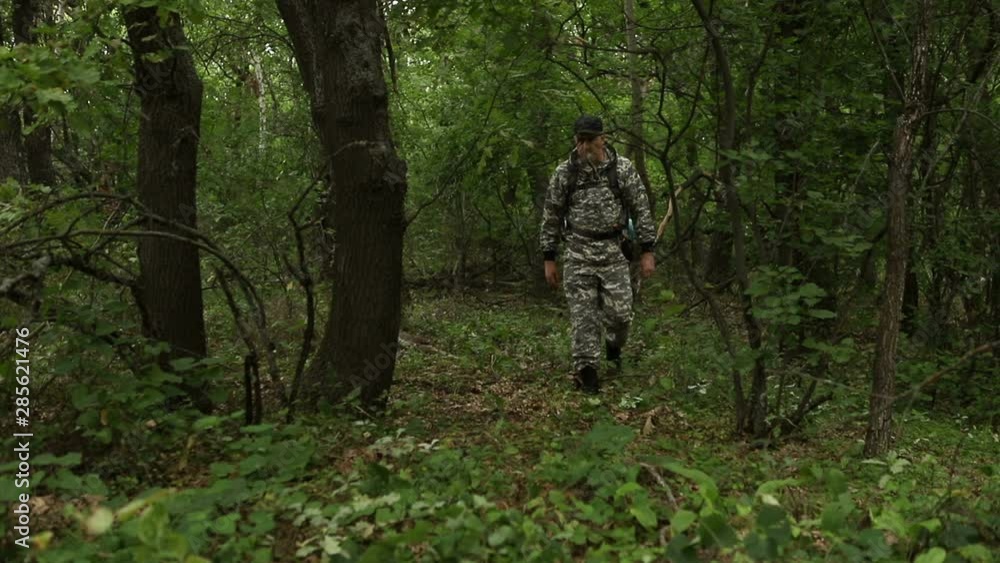 young handsome сaucasian man in a camouflage suit, cap and with a backpack, walks through the dense green forest past the camera, the average plan young handsome сaucasian man in a camouflage suit, cap and with a backpack, walks through the dense green forest past the camera, the average plan
