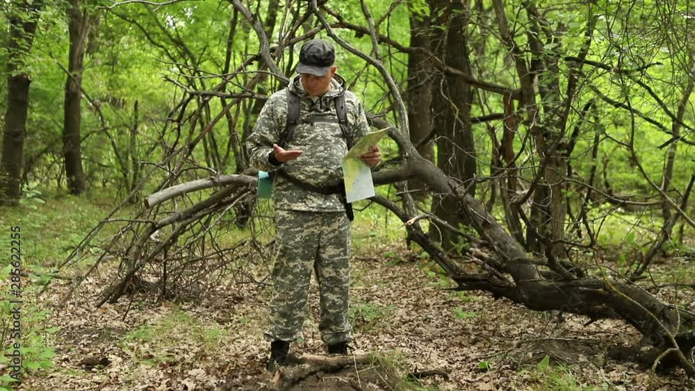 young handsome сaucasian man in a camouflage suit, cap and with a backpack, sits on vacation in a dense green forest and checks his route on a map and a compass, the average plan young handsome сaucasian man in a camouflage suit, cap and with a backpack, sits on vacation in a dense green forest and checks his route on a map and a compass, the average plan