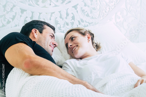 Young happy people caucasian beautiful couple smile and look eachother laying in the bed at home or hotel - travel or real life at ome - bedroom activity in the morning love relationship millennial photo