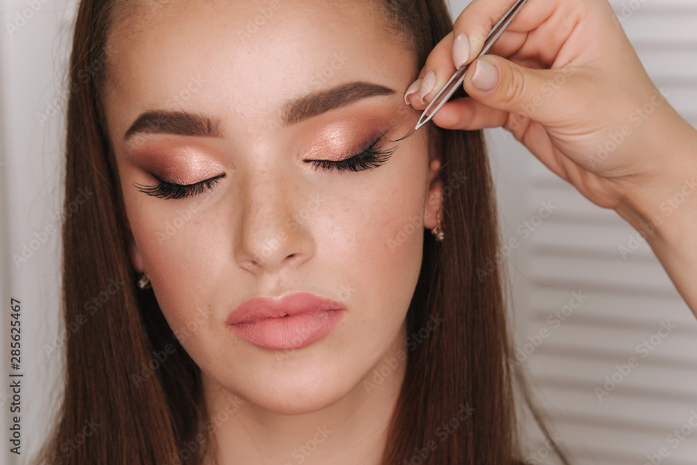 Close up view of make-up artist puts false eyelashes using tweezers. Background of white folding screen Close up view of make-up artist puts false eyelashes using tweezers. Background of white folding screen