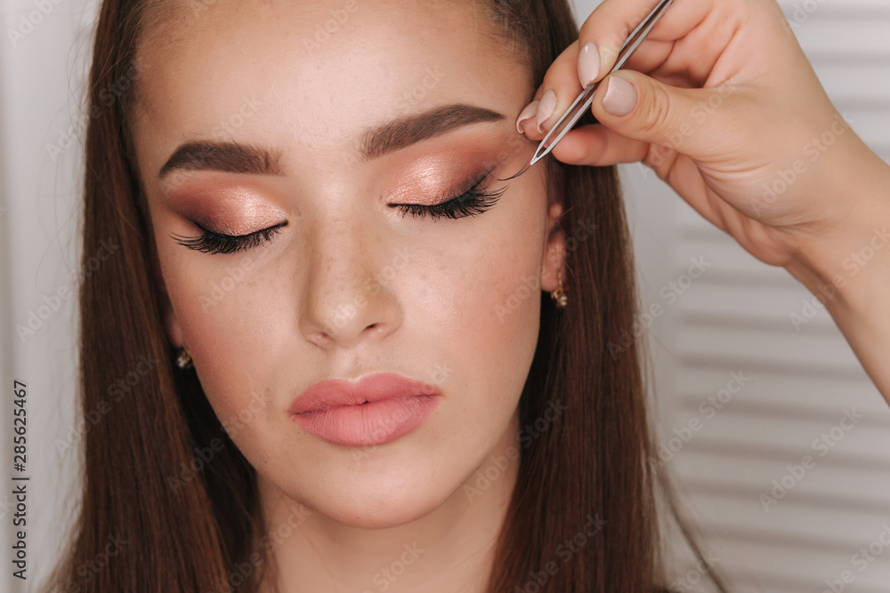 Close up view of make-up artist puts false eyelashes using tweezers. Background of white folding screen