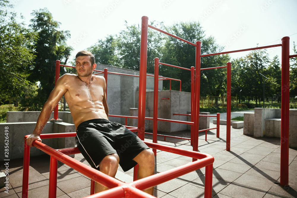 Fototapeta premium Young muscular shirtless caucasian man doing crunches on horizontal bar at playground in sunny summer's day. Training upper body outdoors. Concept of sport, workout, healthy lifestyle, wellbeing.