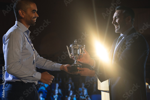 Businessman giving trophy to business male executive on stage in auditorium photo