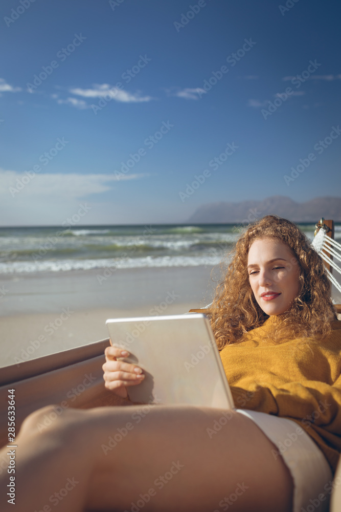 Fototapeta premium Woman using digital tablet while lying on hammock at beach 