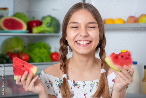 Beautiful young teen girl holding slice ripe red juicy watermelon while standing near open fridge in kitchen at home. Portrait of pretty child choosing food in refrigerator full of healthy products.