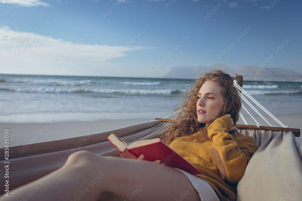 Woman holding book while lying on hammock at beach 