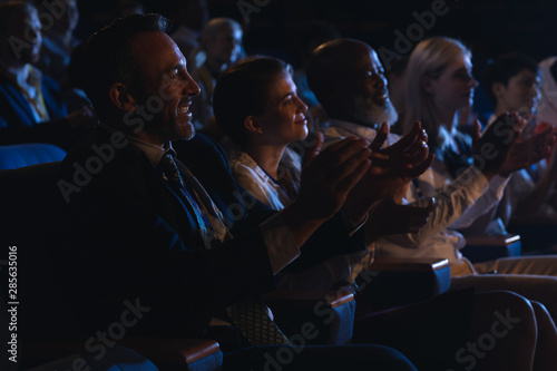 Business colleagues sitting and watching presentation with audience and clapping hands 