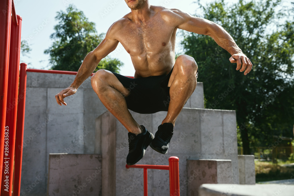 Young muscular shirtless caucasian man jumping above horizontal bar at playground in sunny summer's day. Training his upper body outdoors. Concept of sport, workout, healthy lifestyle, wellbeing.