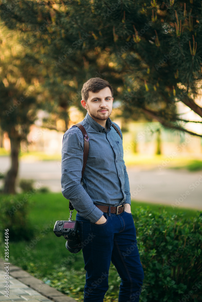 Portrait of professional photographer outside. Man use best camera and leather strap. Handsome confident young man in shirt, standing against street