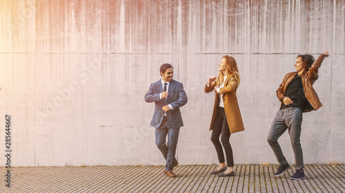 Cheerful Girl and Happy Young Man with Long Hair are Actively Dancing on a Street. Businessman in a Suit Joins Them. They Wear Brown Leather Jacket and Coat. Sunny Day.