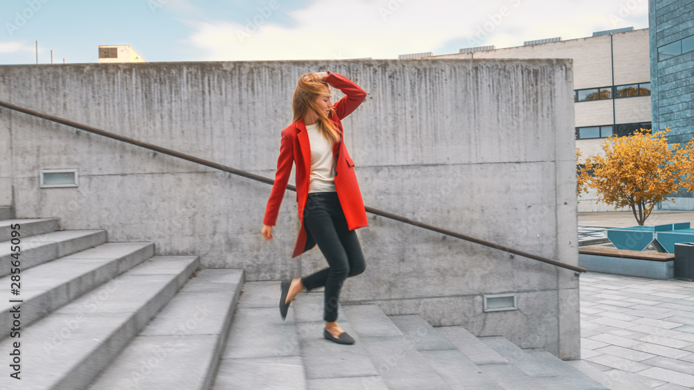 Cheerful and Happy Young Woman Actively Dancing While Walking Down the Stairs. She's Wearing a Long Red Coat. Scene Shot in an Urban Concrete Park Next to Business Center. Day is Bright.