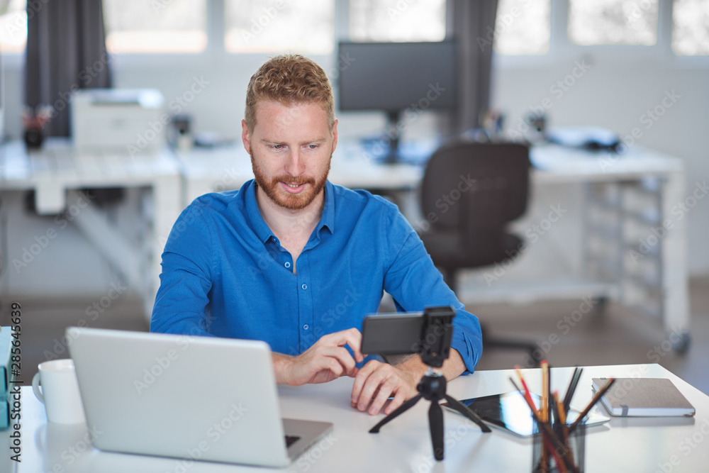 Fototapeta premium Young Caucasian employee having a video call over smart phone while sitting in office. On desk is laptop. Start up business concept.