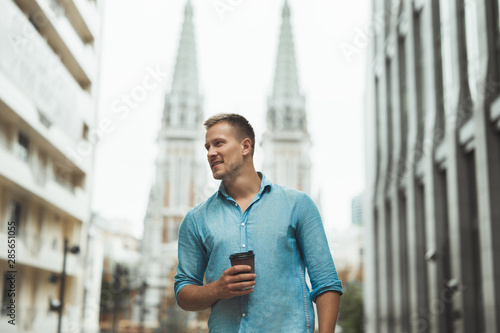 young handsome man holding cup of coffee walking down the street downtown feeling happy