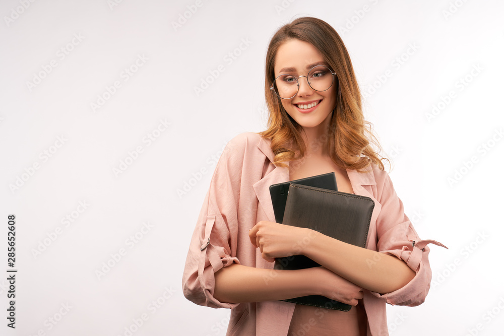 Close up photo of young pretty long haired university teacher, dressed in pink romantic clothes, holding file folder and ipad in her hands, looking a bit down with beautiful embarrassed smile. Close up photo of young pretty long haired university teacher, dressed in pink romantic clothes, holding file folder and ipad in her hands, looking a bit down with beautiful embarrassed smile.