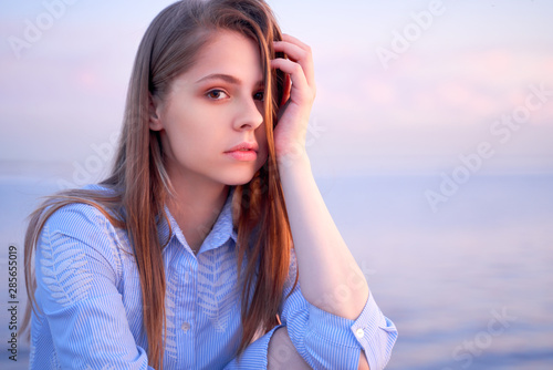 Close up portrait of lovely dark haired teenage girl, dressed in casual summer dress, looking straight at camera with pleasant tender and pensive look, holding hand on her head, posing outdoors. 