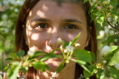 A young smiling girl with dark brown eyes stands in a blooming Apple tree in the spring and sniffs the flowers on a pleasant Sunny day. Large portrait of a beautiful woman in the Park © EverGrump