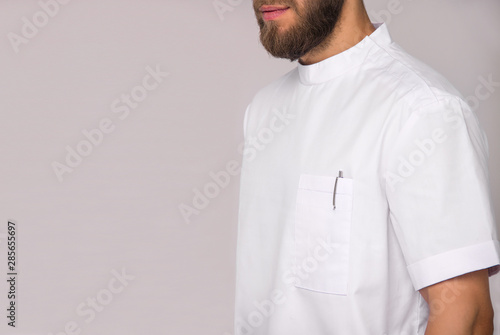 close-up of young stylish bearded nurse man in white medical shirt with pen in pocket which is standing and smiling on white wall background. medical fashion concept. free space on left side