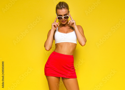 young sports happy sexy blonde girl in red skirt and white top is posing like model and looking down with sunglasses in her hands on the yellow wall background, sport lifestyle concept, free space