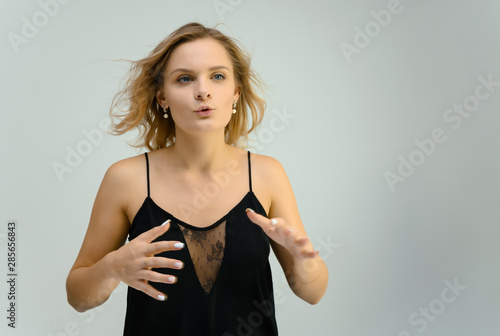 Photo studio portrait of a cute blonde young woman girl in a black blouse on a white background. He stands right in front of the camera  explains with emotion.