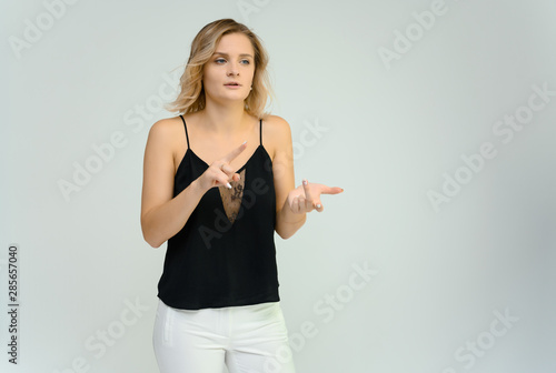 Photo studio portrait of a cute blonde young woman girl in a black blouse and white pants on a white background. He stands right in front of the camera, explains with emotion.