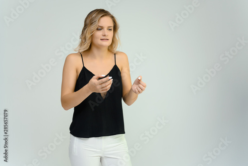 Photo studio portrait of a cute blonde young woman girl in a black blouse and white pants on a white background. He stands right in front of the camera, explains with emotion.