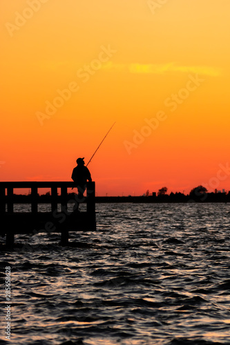 Silhouette of a fisherman looking off into the distance across the ocean, as the sunset lights up the sky with an orange glow. Jones Beach State Park Fishing Piers, Long Island New York. 