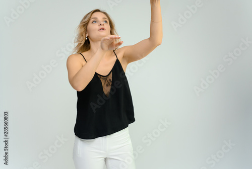 Photo studio portrait of a cute blonde young woman girl in a black blouse and white pants on a white background. He stands right in front of the camera  explains with emotion.