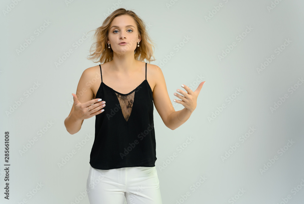 Photo studio portrait of a cute blonde young woman girl in a black blouse and white pants on a white background. He stands right in front of the camera, explains with emotion. Photo studio portrait of a cute blonde young woman girl in a black blouse and white pants on a white background. He stands right in front of the camera, explains with emotion.