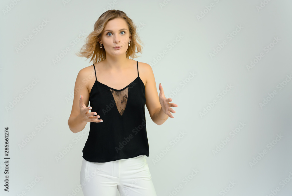 Photo studio portrait of a cute blonde young woman girl in a black blouse and white pants on a white background. He stands right in front of the camera, explains with emotion. Photo studio portrait of a cute blonde young woman girl in a black blouse and white pants on a white background. He stands right in front of the camera, explains with emotion.