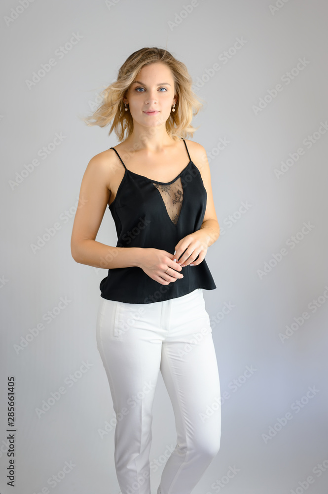 Full length studio portrait photo of a cute young blonde woman girl in a black blouse and white pants on a white background. He stands right in front of the camera, explains with emotion. Full length studio portrait photo of a cute young blonde woman girl in a black blouse and white pants on a white background. He stands right in front of the camera, explains with emotion.