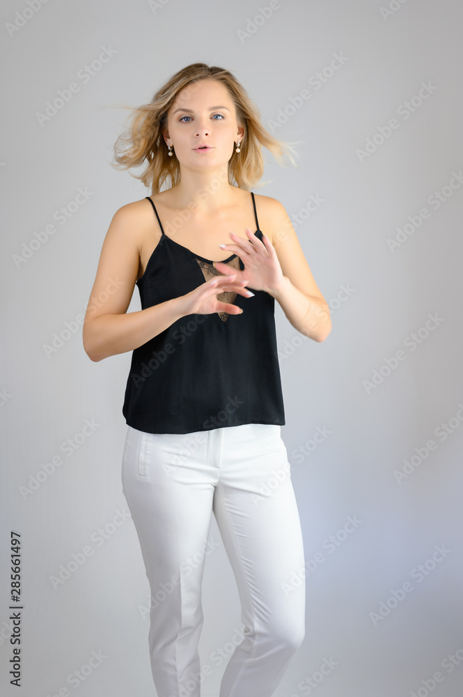 Full length studio portrait photo of a cute young blonde woman girl in a black blouse and white pants on a white background. He stands right in front of the camera, explains with emotion. Full length studio portrait photo of a cute young blonde woman girl in a black blouse and white pants on a white background. He stands right in front of the camera, explains with emotion.