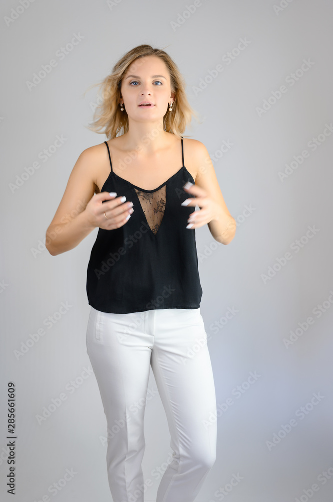 Full length studio portrait photo of a cute young blonde woman girl in a black blouse and white pants on a white background. He stands right in front of the camera, explains with emotion. Full length studio portrait photo of a cute young blonde woman girl in a black blouse and white pants on a white background. He stands right in front of the camera, explains with emotion.