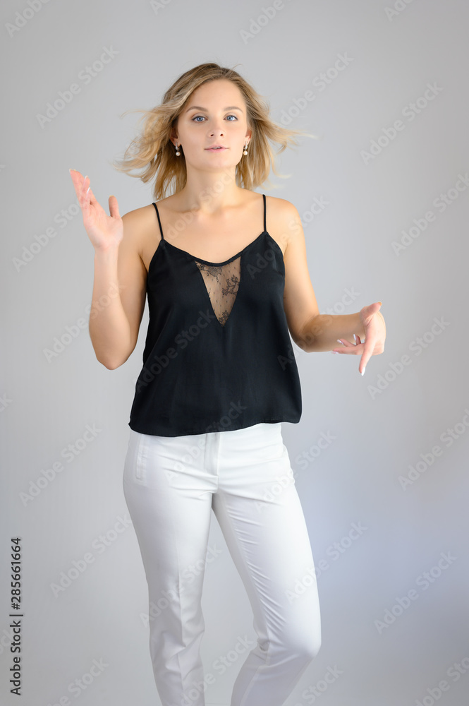 Full length studio portrait photo of a cute young blonde woman girl in a black blouse and white pants on a white background. He stands right in front of the camera, explains with emotion. Full length studio portrait photo of a cute young blonde woman girl in a black blouse and white pants on a white background. He stands right in front of the camera, explains with emotion.