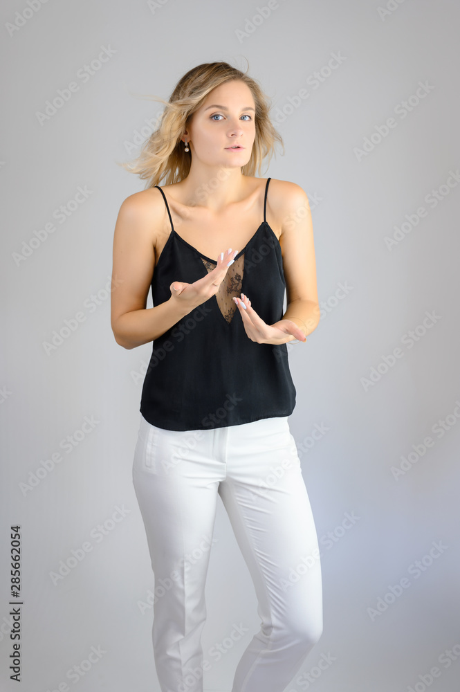 Full length studio portrait photo of a cute young blonde woman girl in a black blouse and white pants on a white background. He stands right in front of the camera, explains with emotion. Full length studio portrait photo of a cute young blonde woman girl in a black blouse and white pants on a white background. He stands right in front of the camera, explains with emotion.