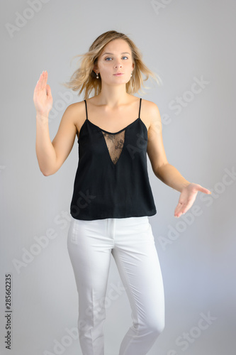 Full length studio portrait photo of a cute young blonde woman girl in a black blouse and white pants on a white background. He stands right in front of the camera  explains with emotion.