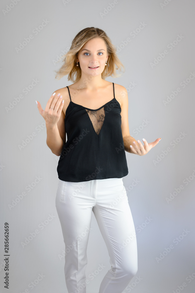 Full length studio portrait photo of a cute young blonde woman girl in a black blouse and white pants on a white background. He stands right in front of the camera, explains with emotion. Full length studio portrait photo of a cute young blonde woman girl in a black blouse and white pants on a white background. He stands right in front of the camera, explains with emotion.