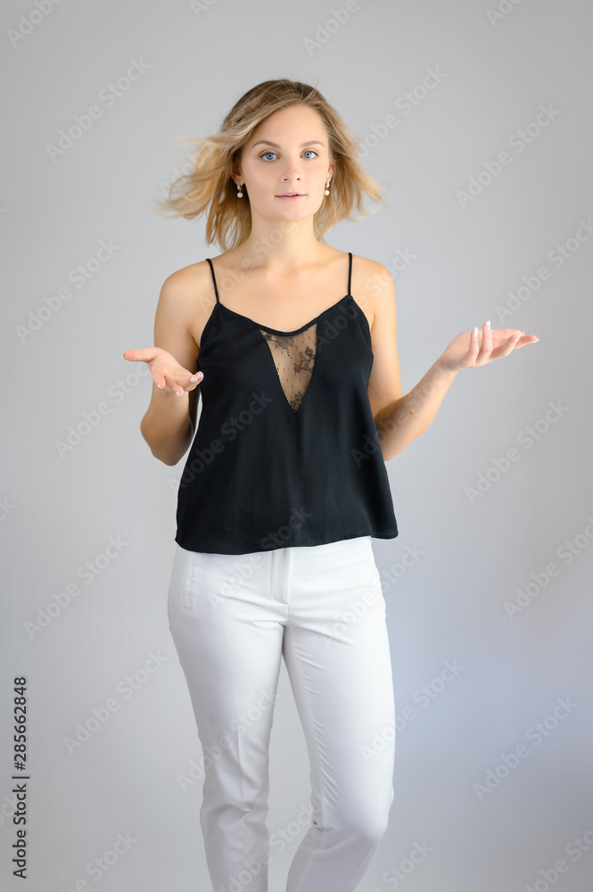 Full length studio portrait photo of a cute young blonde woman girl in a black blouse and white pants on a white background. He stands right in front of the camera, explains with emotion. Full length studio portrait photo of a cute young blonde woman girl in a black blouse and white pants on a white background. He stands right in front of the camera, explains with emotion.