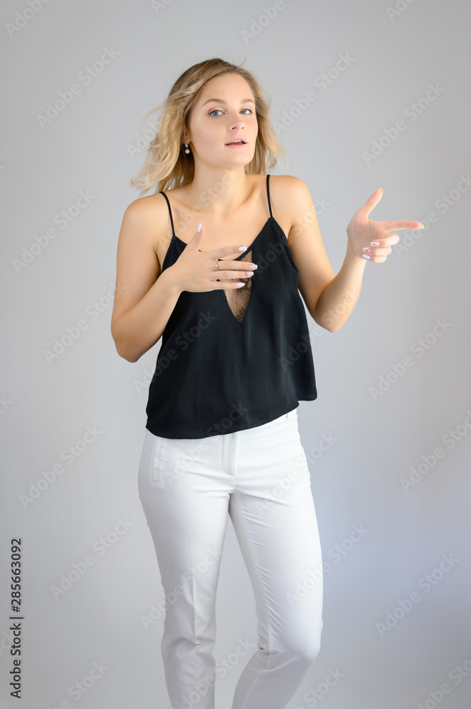 Full length studio portrait photo of a cute young blonde woman girl in a black blouse and white pants on a white background. He stands right in front of the camera, explains with emotion. Full length studio portrait photo of a cute young blonde woman girl in a black blouse and white pants on a white background. He stands right in front of the camera, explains with emotion.