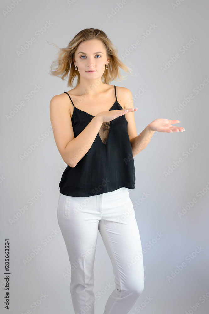 Full length studio portrait photo of a cute young blonde woman girl in a black blouse and white pants on a white background. He stands right in front of the camera, explains with emotion. Full length studio portrait photo of a cute young blonde woman girl in a black blouse and white pants on a white background. He stands right in front of the camera, explains with emotion.