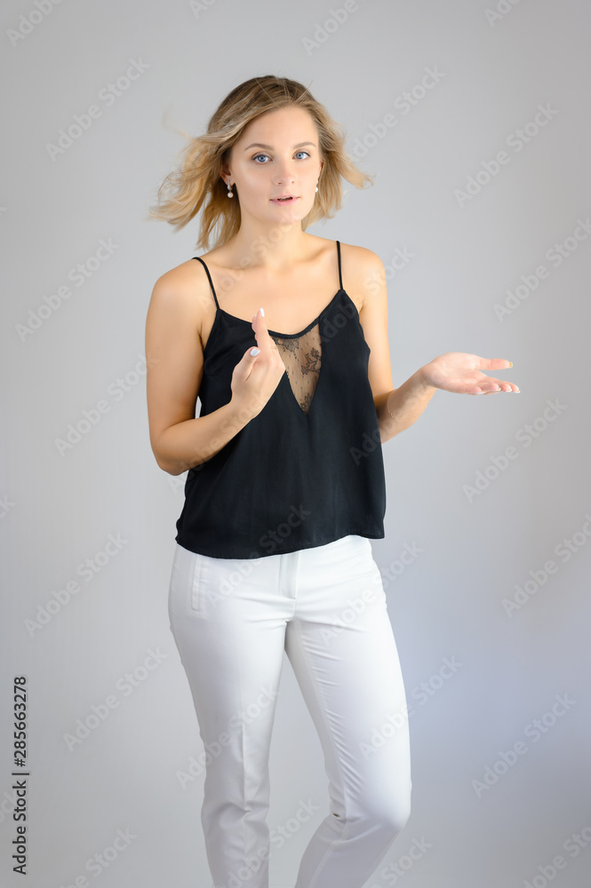 Full length studio portrait photo of a cute young blonde woman girl in a black blouse and white pants on a white background. He stands right in front of the camera, explains with emotion. Full length studio portrait photo of a cute young blonde woman girl in a black blouse and white pants on a white background. He stands right in front of the camera, explains with emotion.