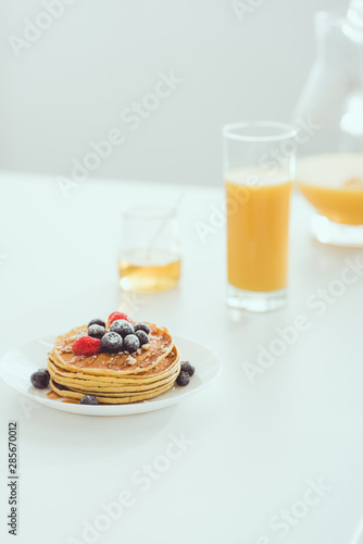 selective focus of plate with tasty pancakes and berries near glass and jug of orange juice and jar with honey on white table