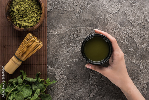 top view of woman holding matcha tea on table with whisk, tea powder and mint