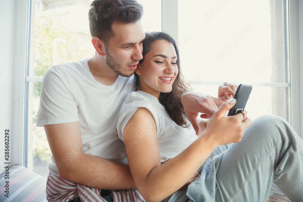Let's catch this moment. Young couple moved to a new house or apartment. Look happy and confident. Family, moving, relations, first home concept. Sitting near by window, hugging and making selfie. Let's catch this moment. Young couple moved to a new house or apartment. Look happy and confident. Family, moving, relations, first home concept. Sitting near by window, hugging and making selfie.