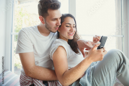 Let's catch this moment. Young couple moved to a new house or apartment. Look happy and confident. Family, moving, relations, first home concept. Sitting near by window, hugging and making selfie.