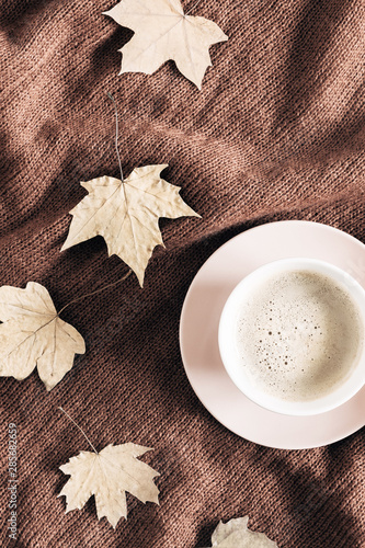 Autumn home cozy composition. Cup of coffee, knitted plaid, dried autumn leaves on brown background. Fall background. Flat lay, top view, copy space