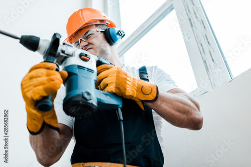 low angle view of handyman in helmet and yellow gloves using hammer drill