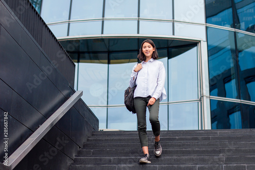Canvas Print Happy cheerful asian student girl going down the stairs from modern office business centre with backpack in white shirt, outside