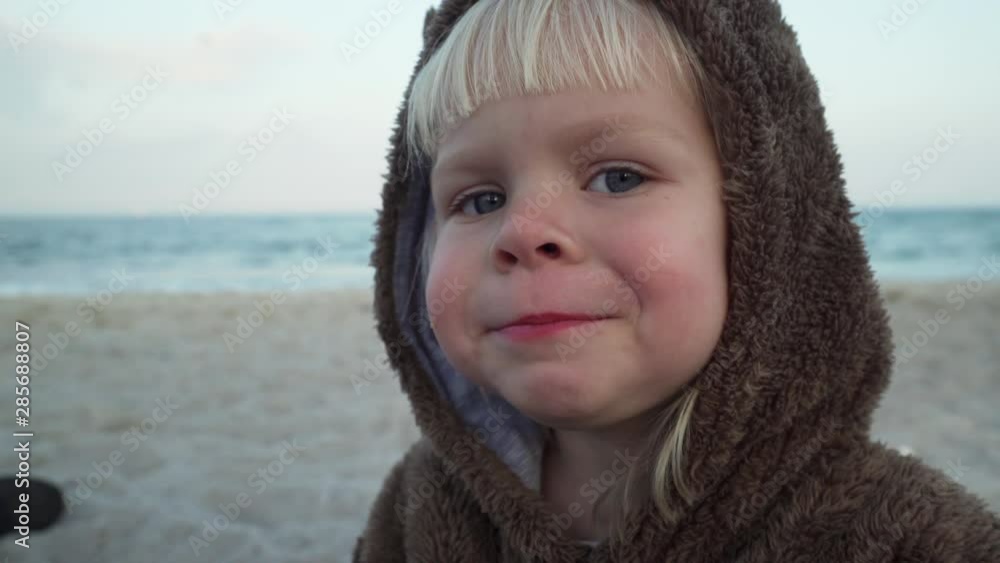 Blonde baby girl in bear hoodie chew a pastry at sandy sea shore cloudy weather Blonde baby girl in bear hoodie chew a pastry at sandy sea shore cloudy weather